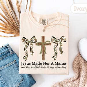 May include: Ivory-colored t-shirt with a graphic featuring a wooden cross and two camouflage bows. The text "Jesus Made Her A Mama and she wouldn't have it any other way" is printed below the graphic. The shirt is laid flat with a pair of blue jeans and white shoes.