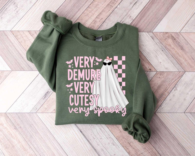 May include: A green sweatshirt with a pink and white graphic of a ghost wearing sunglasses and a bow. The text on the graphic reads "Very Demure Very Cutesy Very Spooky".