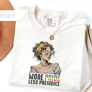 May include: Ivory-colored t-shirt featuring a graphic of a woman wearing heart-shaped rainbow sunglasses and a floral crown. The text "MORE PRIDE LESS PREJUDICE" is printed below the graphic. A gold necklace is draped on the shirt.