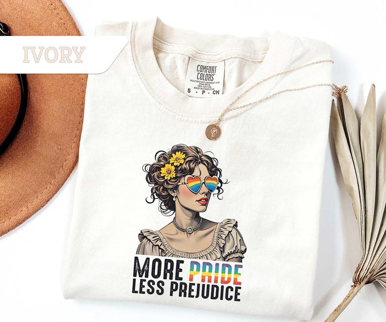 May include: Ivory-colored t-shirt featuring a graphic of a woman wearing heart-shaped rainbow sunglasses and a floral crown. The text "MORE PRIDE LESS PREJUDICE" is printed below the graphic. A gold necklace is draped on the shirt.