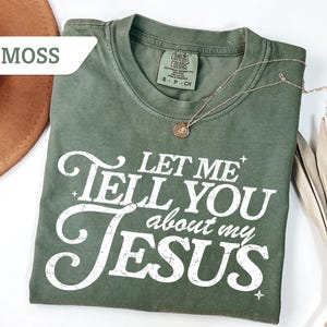 Puede incluir: Una camiseta verde oliva con el texto "LET ME TELL YOU about my JESUS" en blanco. Un collar dorado con un colgante est&aacute; drapeado sobre la camisa. La camisa est&aacute; doblada sobre una superficie blanca.