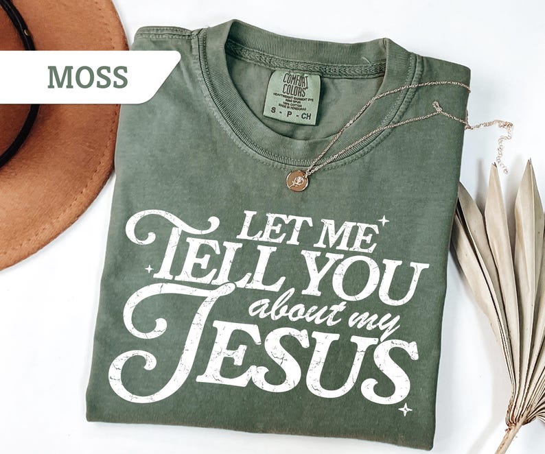 Puede incluir: Una camiseta verde oliva con el texto "LET ME TELL YOU about my JESUS" en blanco. Un collar dorado con un colgante est&aacute; drapeado sobre la camisa. La camisa est&aacute; doblada sobre una superficie blanca.