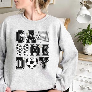Puede incluir: Sudadera gris con el texto "GAME DAY" en letras negras. El texto est&aacute; decorado con un bal&oacute;n de f&uacute;tbol, una porter&iacute;a de f&uacute;tbol y un lazo con un patr&oacute;n de bal&oacute;n de f&uacute;tbol.