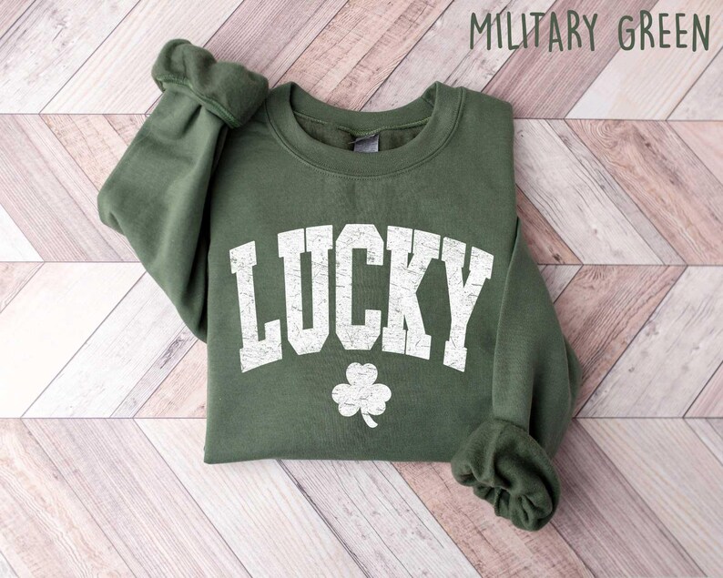 Peut inclure: Un sweat-shirt vert avec le mot "LUCKY" en lettres blanches vieillies et un tr&egrave;fle blanc. Le sweat-shirt est pli&eacute; et pos&eacute; sur une surface en bois.