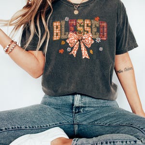 May include: Dark gray t-shirt with the word "BLESSED" in a decorative font. The letters have a plaid, leopard print, and floral design. A bow with a leopard print pattern is below the word, with small flowers and leaves.