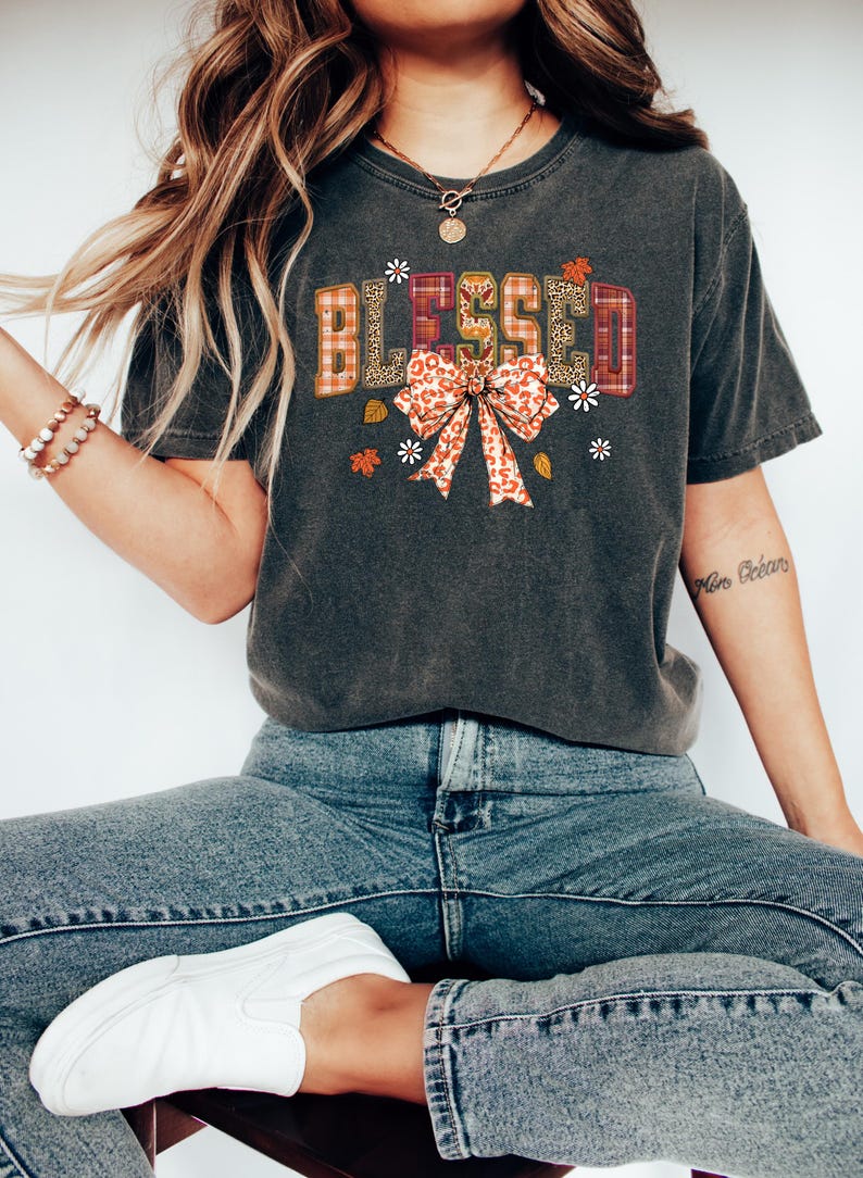 May include: Dark gray t-shirt with the word "BLESSED" in a decorative font. The letters have a plaid, leopard print, and floral design. A bow with a leopard print pattern is below the word, with small flowers and leaves.