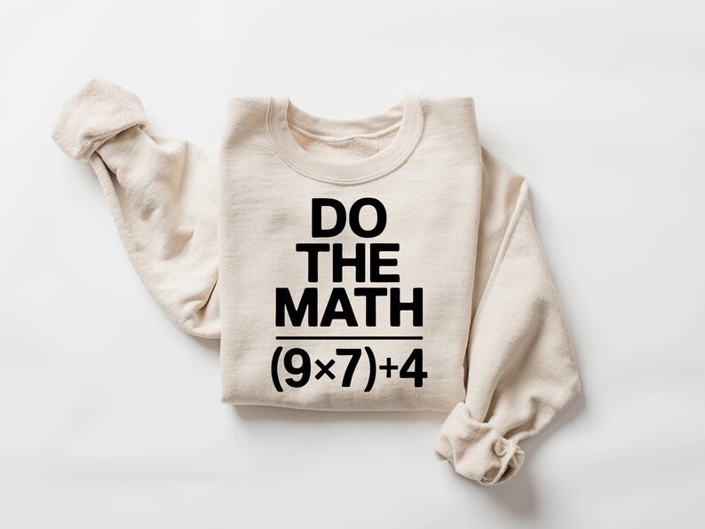 May include: A cream-colored sweatshirt with the text "DO THE MATH" in bold black letters. Below the text is the equation (9x7)+4. The sleeves are rolled up, and the sweatshirt is lying on a white surface.