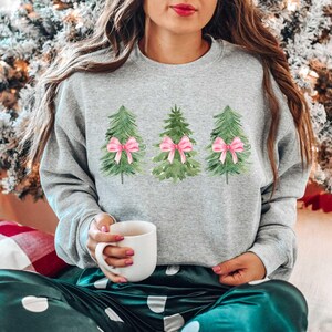 May include: A gray sweatshirt with a watercolor design of three Christmas trees with pink bows. The person wearing the sweatshirt is sitting on a green and white patterned blanket.