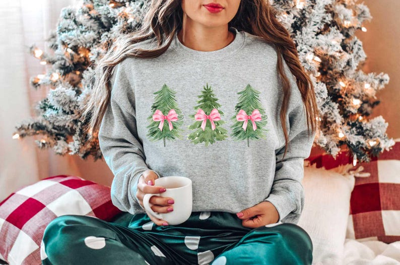 May include: A gray sweatshirt with a watercolor design of three Christmas trees with pink bows. The person wearing the sweatshirt is sitting on a green and white patterned blanket.