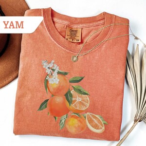 May include: Coral-colored t-shirt with a watercolor illustration of oranges, leaves, and white flowers. A gold necklace with a pendant rests on the shirt. The word "YAM" is in a white banner in the upper left corner. The shirt is folded.