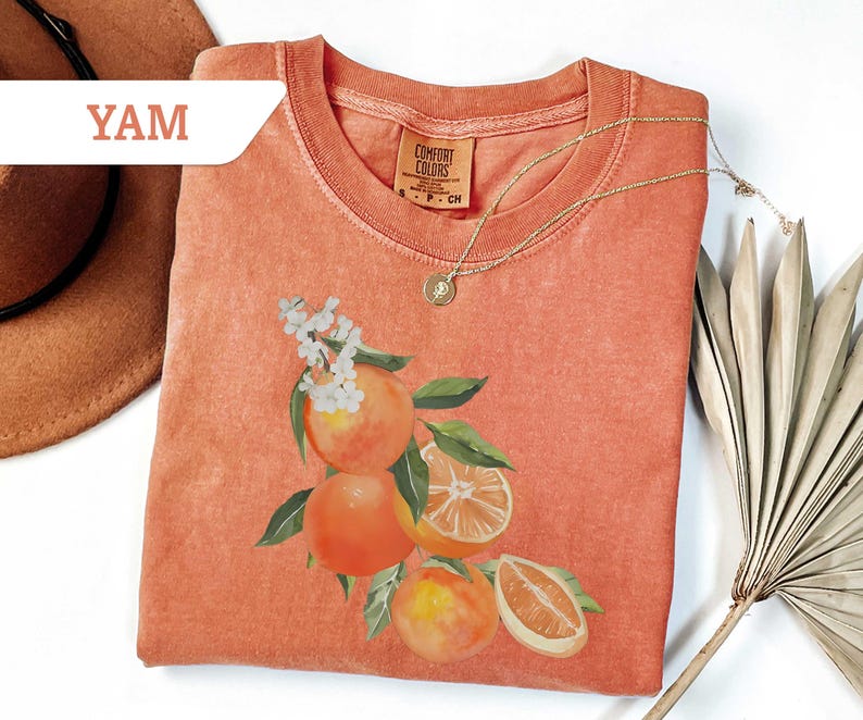 May include: Coral-colored t-shirt with a watercolor illustration of oranges, leaves, and white flowers. A gold necklace with a pendant rests on the shirt. The word "YAM" is in a white banner in the upper left corner. The shirt is folded.