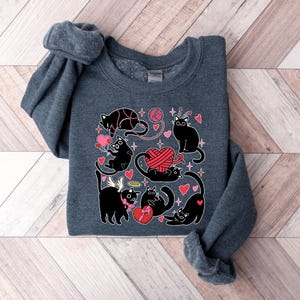 May include: A dark gray sweatshirt with a black cat design. The cats are surrounded by hearts, yarn, and stars. The text "Meow" is printed on the sweatshirt.
