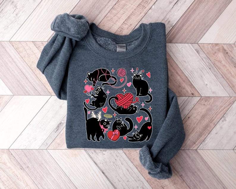 May include: A dark gray sweatshirt with a black cat design. The cats are surrounded by hearts, yarn, and stars. The text "Meow" is printed on the sweatshirt.
