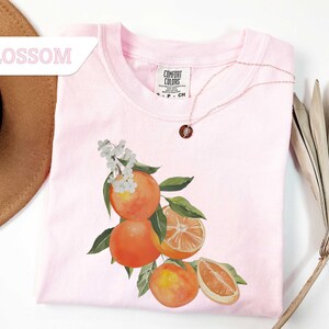 May include: A light pink t-shirt with a watercolor illustration of oranges, leaves, and white blossoms. A delicate gold necklace with a pendant rests on the shirt. The word "BLOSSOM" is in a pink banner in the upper left corner.
