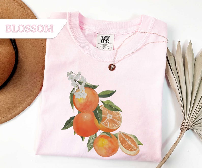 May include: A light pink t-shirt with a watercolor illustration of oranges, leaves, and white blossoms. A delicate gold necklace with a pendant rests on the shirt. The word "BLOSSOM" is in a pink banner in the upper left corner.