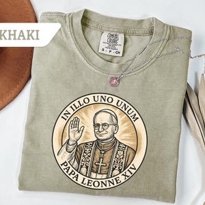 May include: Khaki-colored t-shirt featuring a circular graphic of Pope Leo XIV with the text "IN ILLO UNO UNUM" and "PAPA LEONNE XIV". The shirt has a crew neck and short sleeves. A silver necklace with a pendant is draped across the shirt.