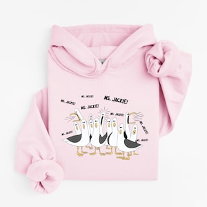 May include: A light pink hoodie featuring a cartoon illustration of seagulls. The seagulls are depicted in black, white, and yellow, with the text "MS. JACKIE!" in black. The hoodie has a drawstring hood and long sleeves.