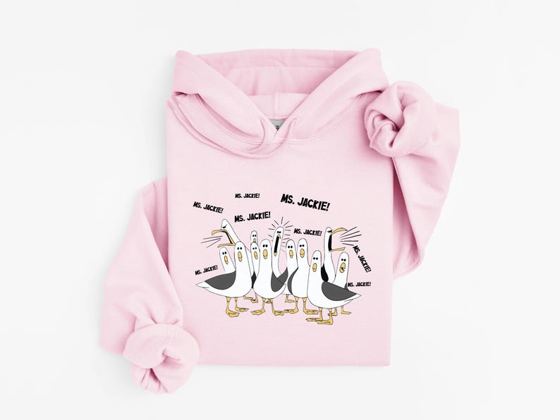 May include: A light pink hoodie featuring a cartoon illustration of seagulls. The seagulls are depicted in black, white, and yellow, with the text "MS. JACKIE!" in black. The hoodie has a drawstring hood and long sleeves.