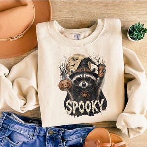 May include: A cream-colored crewneck sweatshirt featuring a graphic of a raccoon wearing a witch hat, holding a pumpkin and a coffee cup, with a full moon and bare trees in the background. The word SPOOKY is printed in a distressed font below.