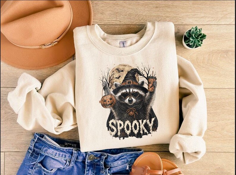 May include: A cream-colored crewneck sweatshirt featuring a graphic of a raccoon wearing a witch hat, holding a pumpkin and a coffee cup, with a full moon and bare trees in the background. The word SPOOKY is printed in a distressed font below.