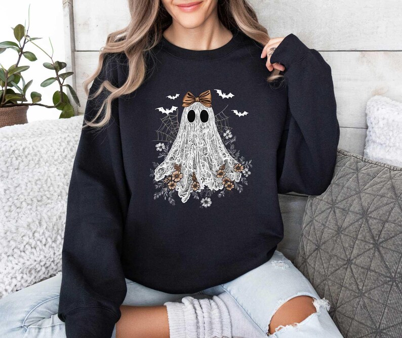 Peut inclure: Sweat-shirt noir avec un fant&ocirc;me blanc portant un n&oelig;ud brun. Le fant&ocirc;me est entour&eacute; de d&eacute;tails floraux et de chauves-souris.