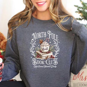 May include: A gray sweatshirt with a white graphic design that says "North Pole Book Club" with a mug of hot cocoa, a gingerbread man, and a stack of books. The text "Hot Cocoa Served Daily" is below the graphic.