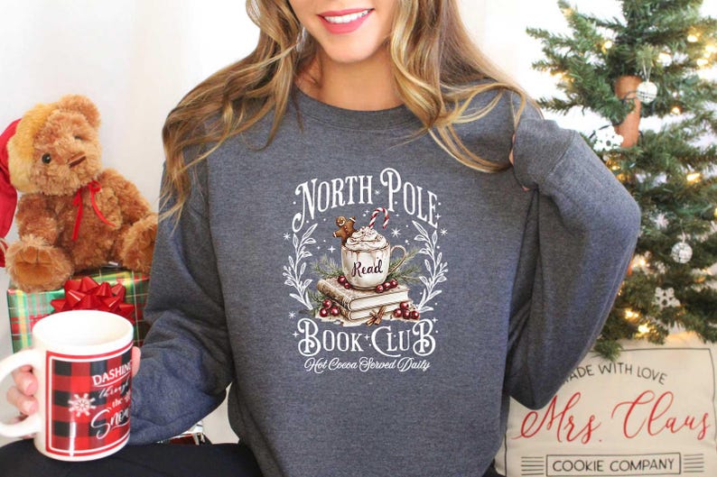 May include: A gray sweatshirt with a white graphic design that says "North Pole Book Club" with a mug of hot cocoa, a gingerbread man, and a stack of books. The text "Hot Cocoa Served Daily" is below the graphic.