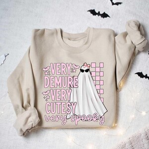 May include: A beige sweatshirt with a pink and white graphic of a ghost with the text "Very Demure Very Cutesy Very Spooky".