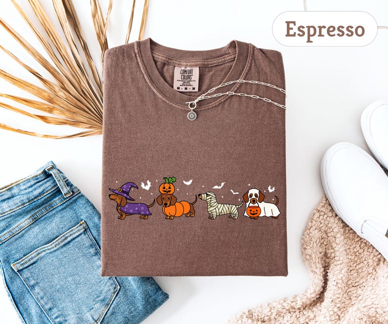 May include: Espresso-colored t-shirt featuring a Halloween-themed dachshund design. The graphic includes dachshunds dressed as a witch, pumpkin, mummy, and holding a jack-o'-lantern. The shirt is paired with blue jeans, white sneakers, and a necklace.