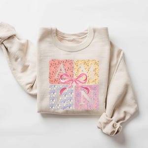 K&ouml;nnte beinhalten: Ein beiger Pullover mit einem Blumendesign und dem Wort "AMYN" in wei&szlig;en Buchstaben mit einer rosa Schleife in der Mitte.