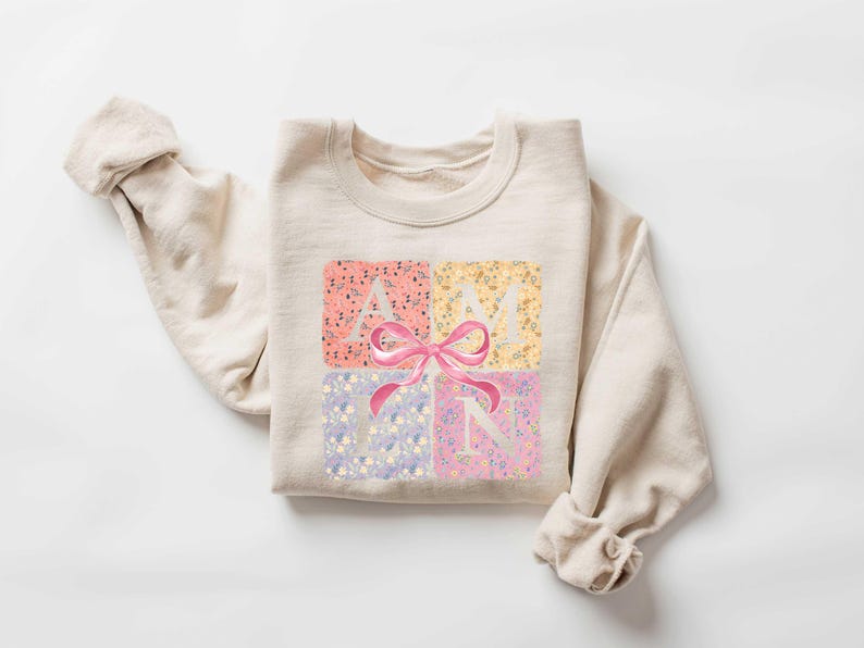 K&ouml;nnte beinhalten: Ein beiger Pullover mit einem Blumendesign und dem Wort "AMYN" in wei&szlig;en Buchstaben mit einer rosa Schleife in der Mitte.