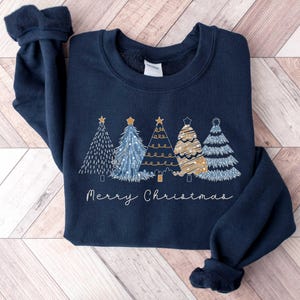 May include: A navy blue sweatshirt featuring a festive design of five stylised Christmas trees in gold and blue with the text "Merry Christmas" underneath.