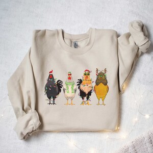 May include: A beige sweatshirt with a Christmas-themed design featuring four chickens wearing Santa hats and reindeer antlers, all tangled in Christmas lights.