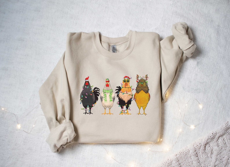 May include: A beige sweatshirt with a Christmas-themed design featuring four chickens wearing Santa hats and reindeer antlers, all tangled in Christmas lights.