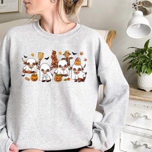 Op de afbeelding: Een grijs sweatshirt met een Halloween-thema ontwerp met een groep cartoon koeien die verkleed zijn als spoken. De koeien houden pompoenen vast en vleermuizen vliegen eromheen.