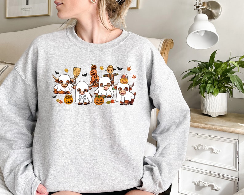 Op de afbeelding: Een grijs sweatshirt met een Halloween-thema ontwerp met een groep cartoon koeien die verkleed zijn als spoken. De koeien houden pompoenen vast en vleermuizen vliegen eromheen.
