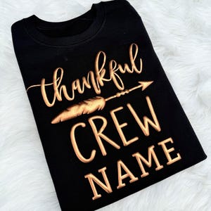 May include: Black crew neck sweatshirt with the words "thankful CREW NAME" in a gold, textured font. A gold feather and arrow design is between the words "thankful" and "CREW". The sweatshirt is folded on a white, fluffy surface.