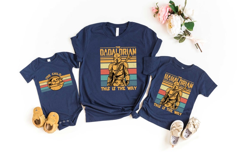 May include: Three navy blue t-shirts with a retro-style graphic of a Mandalorian character. The shirts have the text "The Child", "The Dadalorian", and "The Mamalorian" with the phrase "This is the way" underneath.