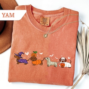 May include: A coral-colored t-shirt featuring a line of dachshund dogs dressed in Halloween costumes. The dogs are wearing a witch hat, pumpkin costume, mummy wrap, and holding a jack-o'-lantern. The shirt has the text "YAM" on the upper left.