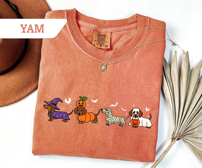 May include: A coral-colored t-shirt featuring a line of dachshund dogs dressed in Halloween costumes. The dogs are wearing a witch hat, pumpkin costume, mummy wrap, and holding a jack-o'-lantern. The shirt has the text "YAM" on the upper left.