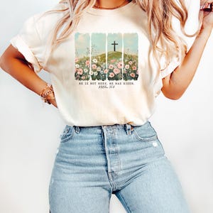 May include: A white t-shirt with a graphic of a cross on a hill with flowers in the foreground. The text "He is not here, He has risen. Matthew 28:6" is printed on the shirt.