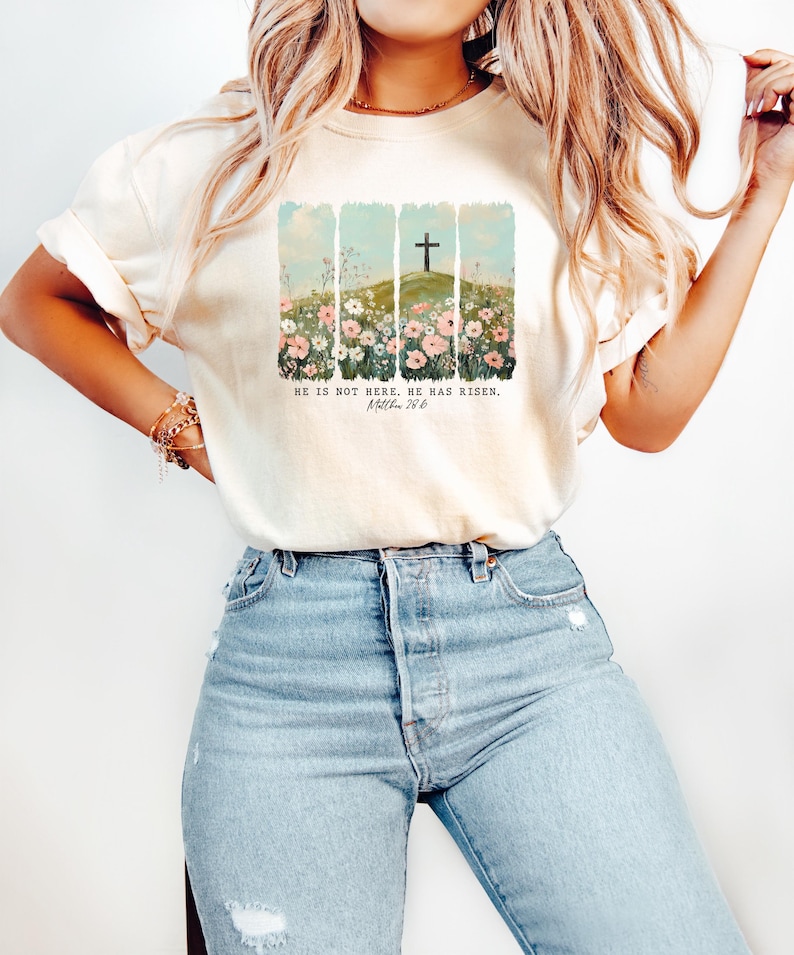 May include: A white t-shirt with a graphic of a cross on a hill with flowers in the foreground. The text "He is not here, He has risen. Matthew 28:6" is printed on the shirt.