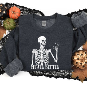 May include: A dark gray sweatshirt with a white skeleton graphic and the text "Never Better". The skeleton is giving a thumbs up. The sweatshirt is surrounded by fall-themed decorations, including pumpkins and leaves.