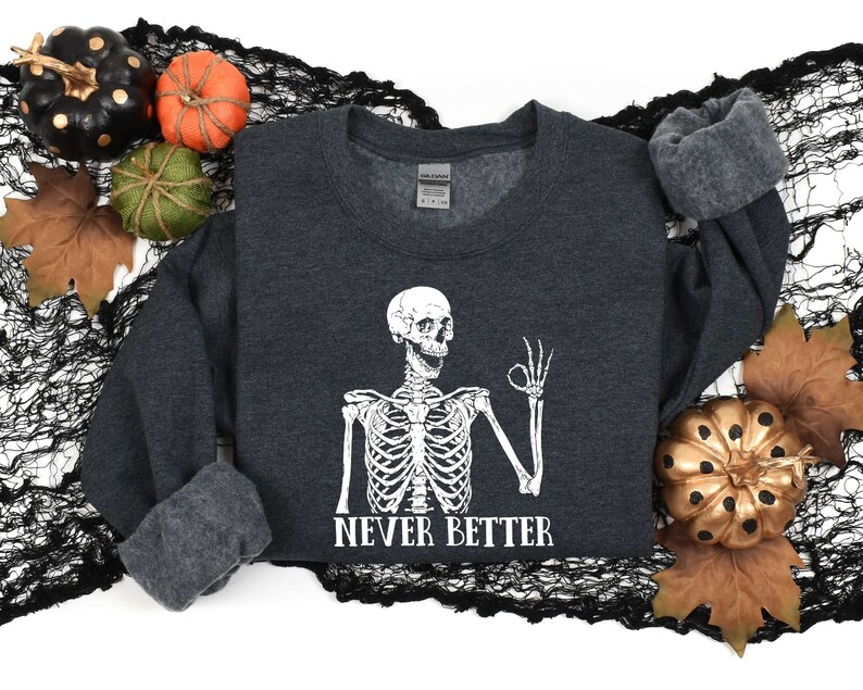 May include: A dark gray sweatshirt with a white skeleton graphic and the text "Never Better". The skeleton is giving a thumbs up. The sweatshirt is surrounded by fall-themed decorations, including pumpkins and leaves.