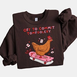 K&ouml;nnte beinhalten: Dunkelbraunes Sweatshirt mit dem Text "OFF TO COMMIT TOMFOOLERY". Ein Cartoon-Huhn mit Zylinder f&auml;hrt auf einem rosa Skateboard mit Herzen. Kleine Herzen und Liebesbriefe sind verstreut.