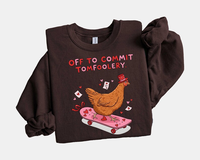 K&ouml;nnte beinhalten: Dunkelbraunes Sweatshirt mit dem Text "OFF TO COMMIT TOMFOOLERY". Ein Cartoon-Huhn mit Zylinder f&auml;hrt auf einem rosa Skateboard mit Herzen. Kleine Herzen und Liebesbriefe sind verstreut.