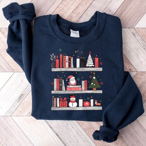 May include: A navy blue sweatshirt with a Christmas-themed design featuring a bookshelf filled with books, a snowman, a Santa Claus, and a Christmas tree. The design is in a white, red, and green colour scheme.
