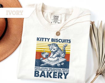 May include: Ivory-colored t-shirt with a graphic that reads "KITTY BISCUITS" above an illustration of a cat kneading dough. The text "PAWS-ITIVELY DELIGHTFUL TREATS WE KNEAD LOVE INTO EVERY BATCH BAKERY" is below the image.