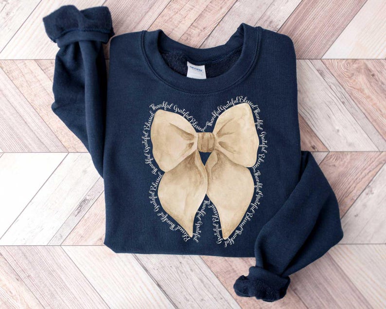 May include: Navy blue sweatshirt with a large beige bow graphic and the text "Thankful Grateful Blessed" repeated around the bow.