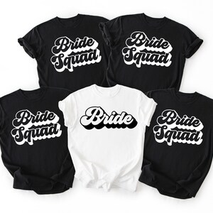 May include: Five t-shirts, four black and one white, with the words "Bride Squad" and "Bride" in a retro font. The black shirts have "Bride Squad" in white. The white shirt has "Bride" in black.